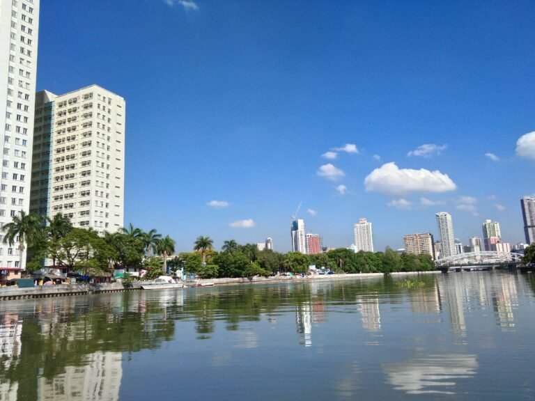 A river runs in the foreground, and in the background, tall buildings stand on the far bank.