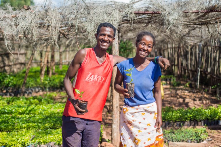 A man and woman pose for the camera in a nursery, smiling and holding small trees.