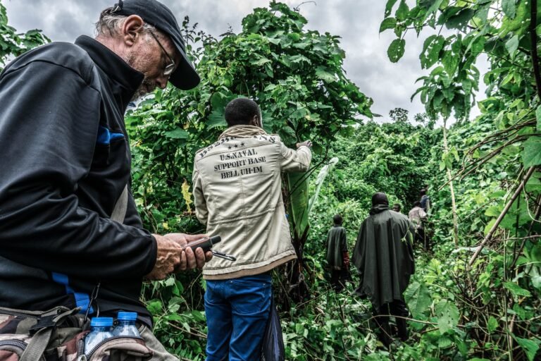A group of people walk on a heavily forested trail, and the man closest to the camera studies a GPS receiver.