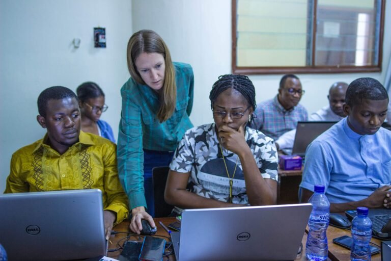 A female trainer assists a group of trainees on computers in a classroom.