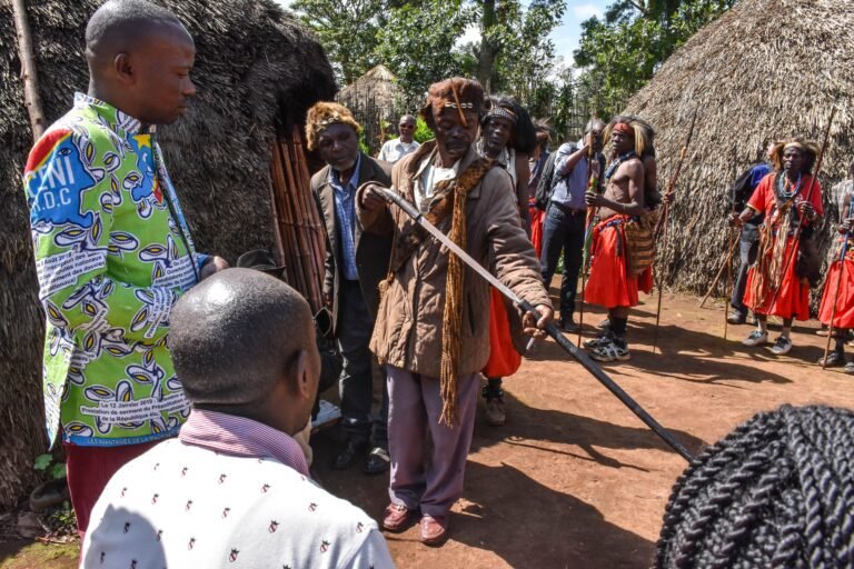 A group of people stand and talk near huts in a village.
