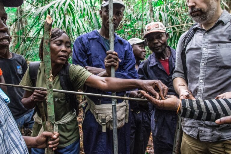 A group of people stand in a forest and hand soil to one another as part of a training exercise in soil measurement.