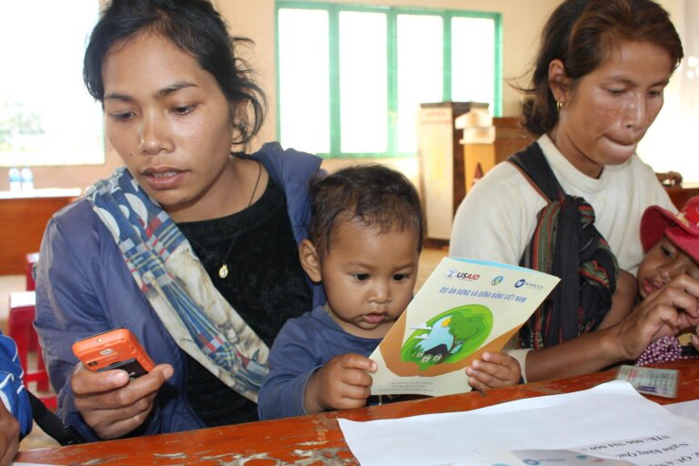 Two women with children in their laps sit at a desk and use their mobile phones to receive money.