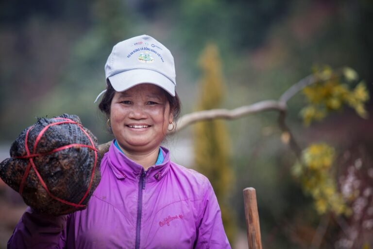 A woman with a young tree on her shoulder smiles at the camera.