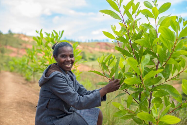 A woman stands next to an acacia tree, holding a branch and smiling at the camera.