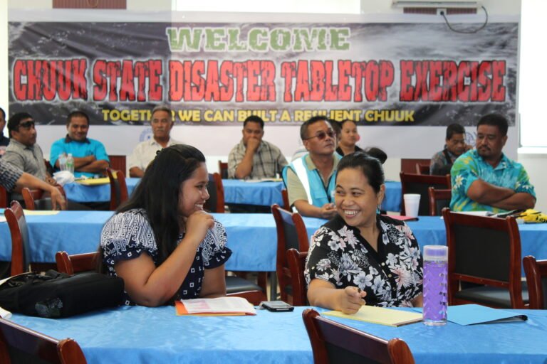 In the foreground, two women sit talking and laughing. A sign behind says 'Chuuk State Disaster Tabletop Exercise.'