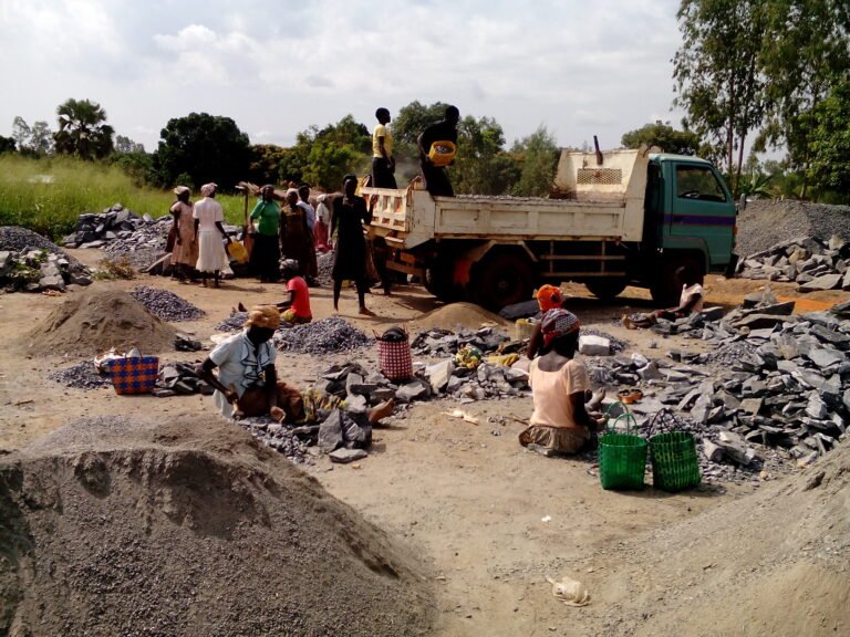 A group of women sits among piles of construction materials while other women help load a truck with some of the materials.