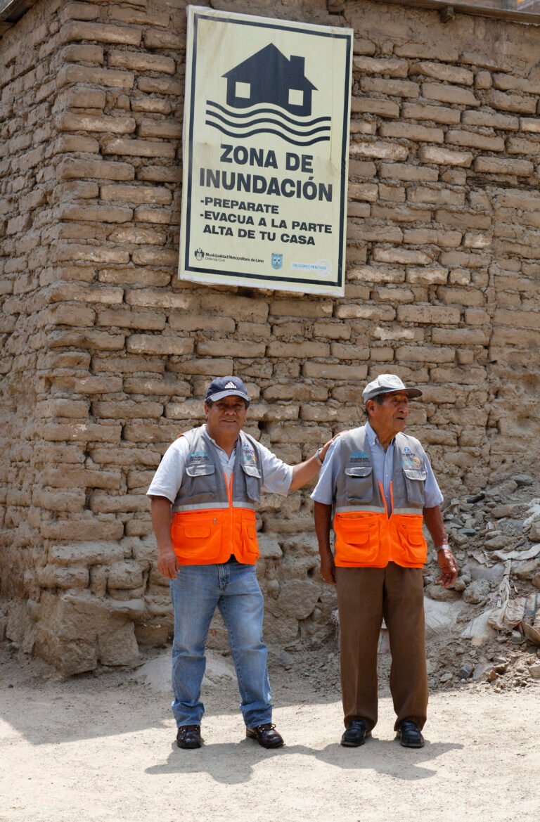 Two construction workers stand in front of a flood zone sign.